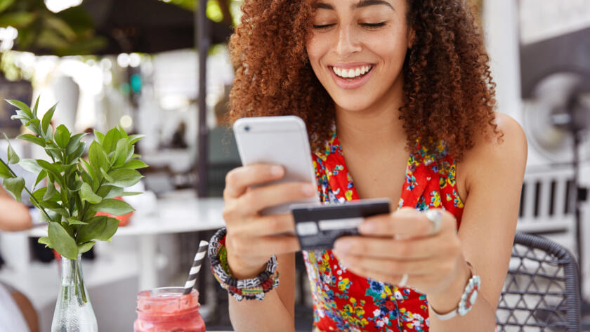 beautiful-dark-skinned-young-female-with-cheerful-expression-holds-smart-phone-credit-card-banks-online-makes-shopping-while-sits-against-cafe-interior-scaled