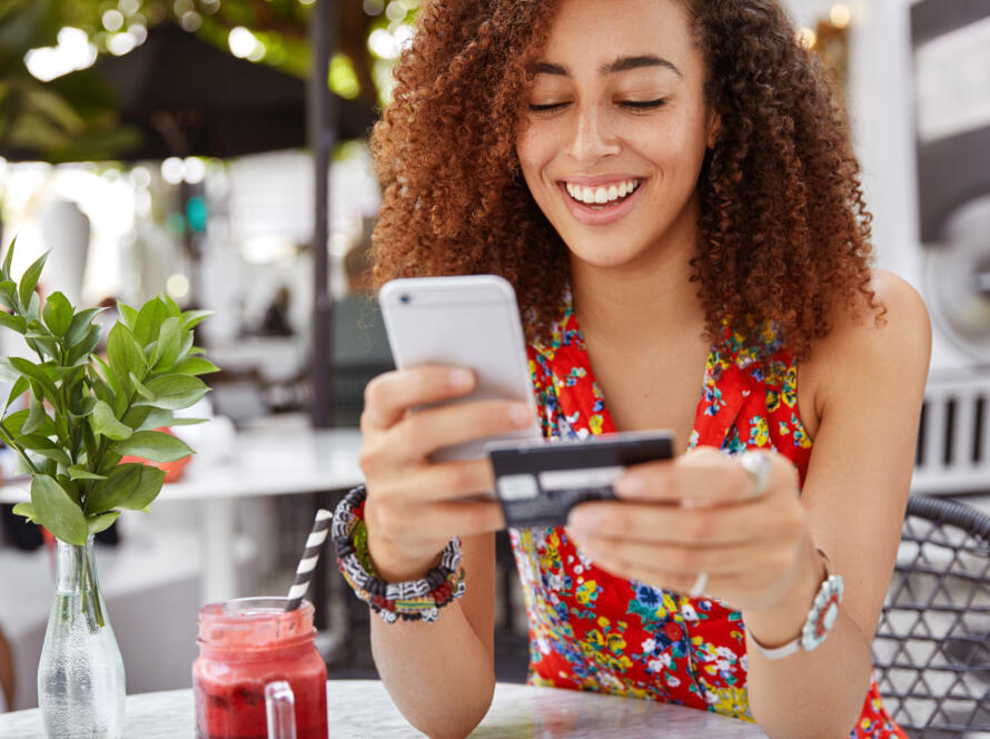 beautiful-dark-skinned-young-female-with-cheerful-expression-holds-smart-phone-credit-card-banks-online-makes-shopping-while-sits-against-cafe-interior-scaled
